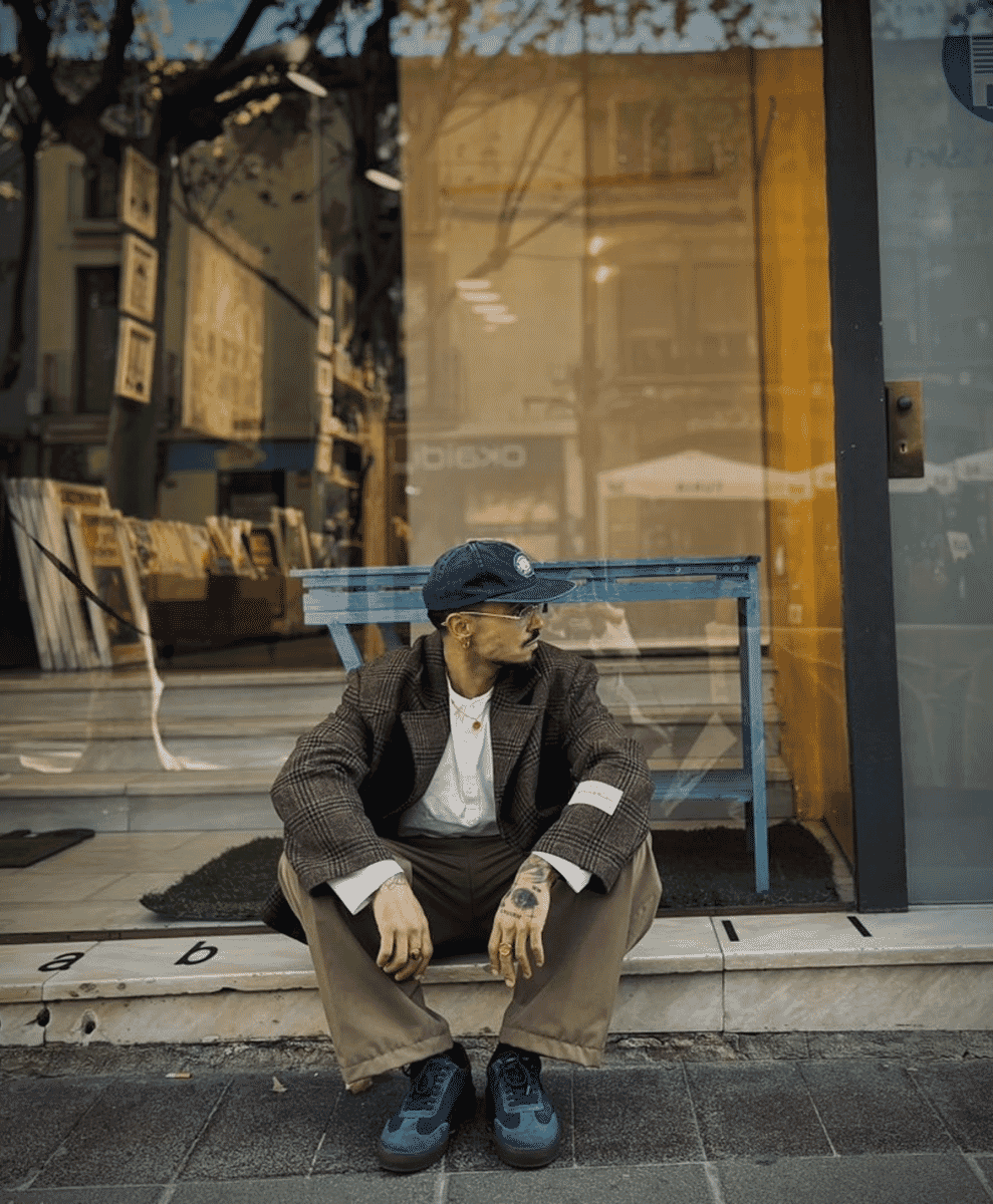 A man in a plaid blazer and blue sneakers sitting on a city sidewalk.
