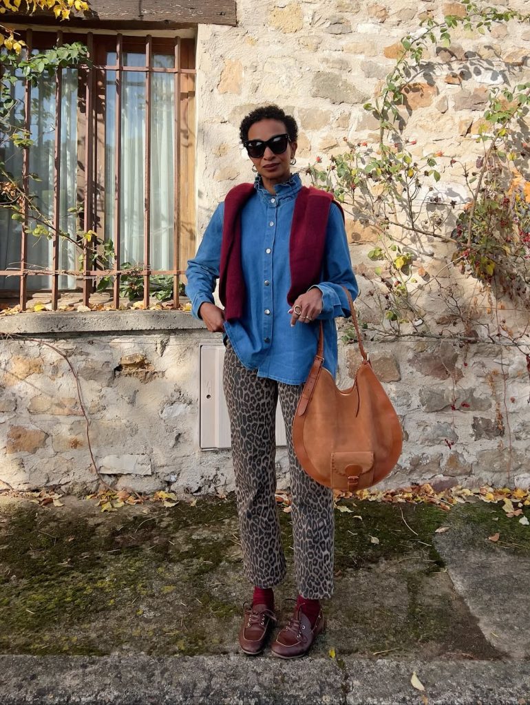 Woman in a denim shirt, red vest, and leopard-print pants carrying a tan leather bag.
