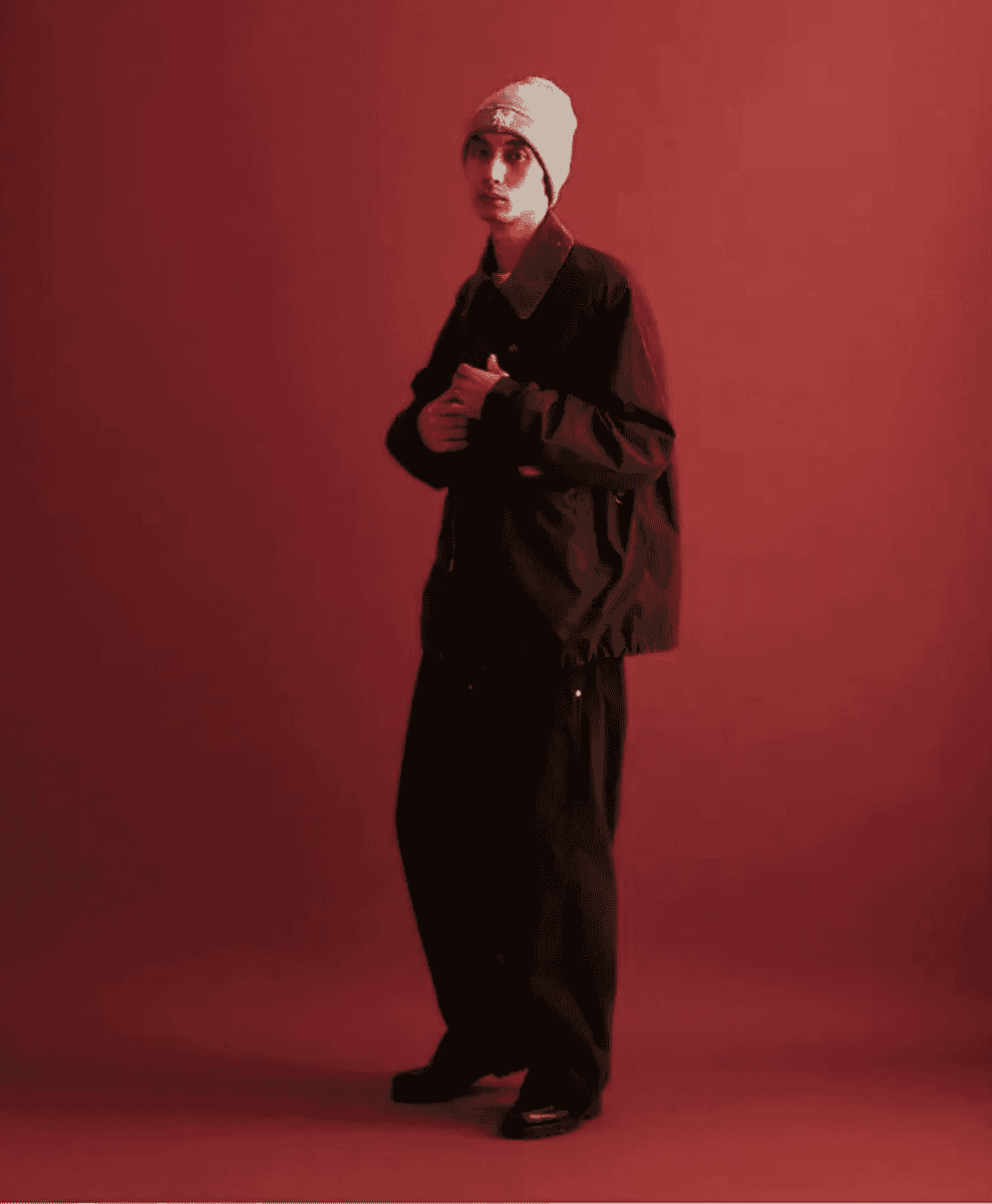 Man in a white beanie and oversized dark jacket against a red background.