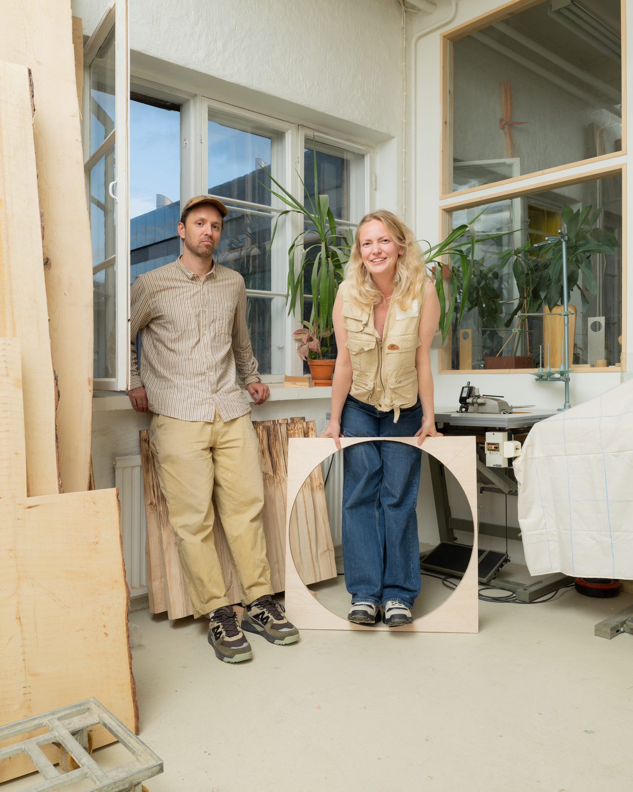 A man and a woman in utility outfits holding a circular wooden panel in a bright, plant-filled workshop.