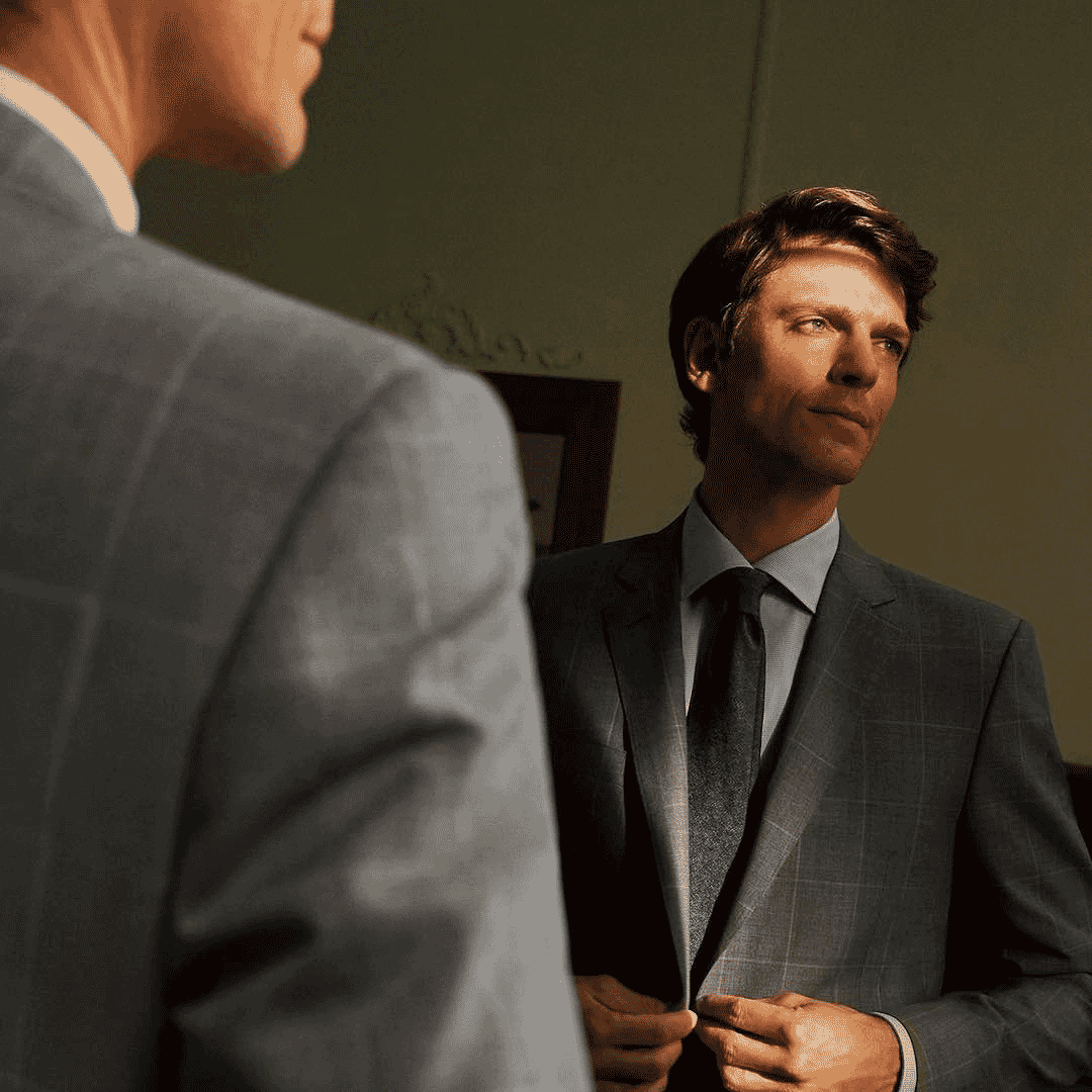 A man in a grey windowpane suit reflecting in a mirror under dramatic lighting.