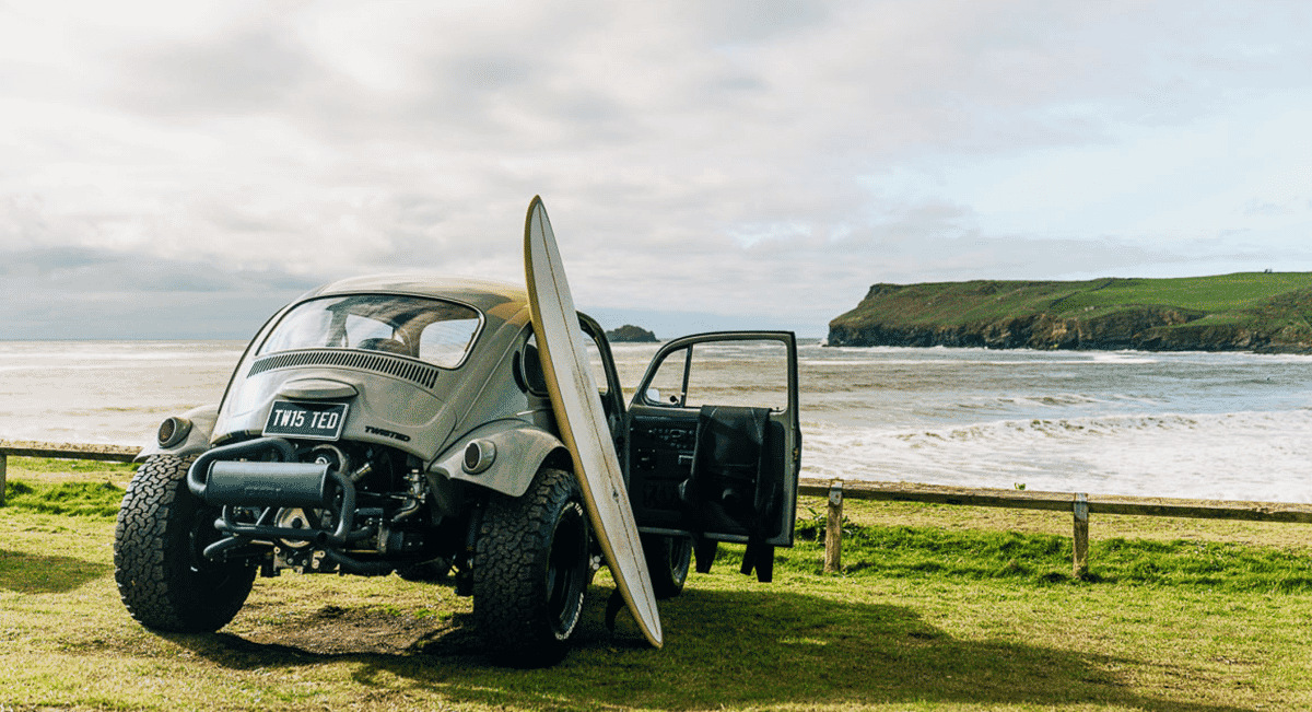 A customized off-road Beetle with a surfboard on top, positioned against a scenic coastal backdrop.