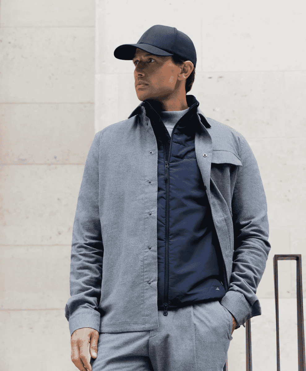 Man in a grey wool coat and navy puffer vest wearing a dark baseball cap against a concrete wall.